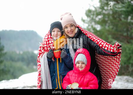 Portrait of family walking in park forest in winter. Happy middle-aged woman mother covering children with blanket. Stock Photo