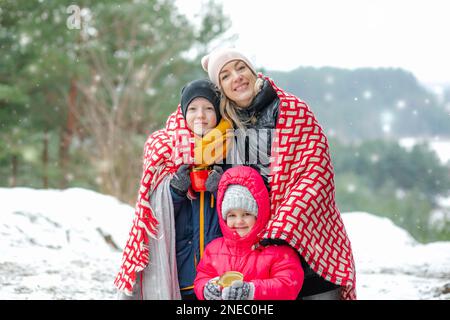 Portrait of family walking in park forest in winter. Middle-aged woman mother covering children with blanket. Snowfall. Stock Photo