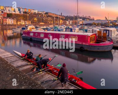 Rowers preparing their boat at sunrise in Bristol Marina, with the colourful terrace houses of Cliftonwood in the distance. Bristol. UK. Stock Photo