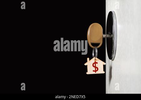 Key with a trinket in the form of a house in the door lock of an open door against the dark background Stock Photo