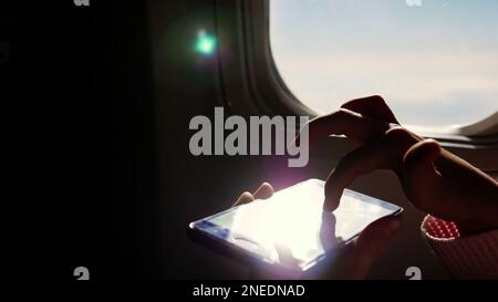 close-up. the sun's rays are reflected in the phone. dark silhouette of kid hands and mobile phone against airplane's illuminator. Child using, plays game on smartphone, tablet phone in airplane cabin. High quality photo Stock Photo