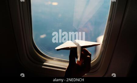 close-up. Silhouette of a child's hand with small paper plane against the background of airplane window. Child sitting by aircraft window and playing with little paper plane. during flight on airplane. High quality photo Stock Photo