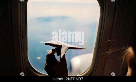 close-up. Silhouette of a child's hand with small paper plane against the background of airplane window. Child sitting by aircraft window and playing with little paper plane. during flight on airplane. High quality photo Stock Photo