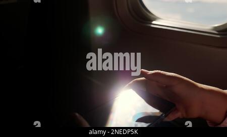 close-up. the sun's rays are reflected in the phone. dark silhouette of kid hands and mobile phone against airplane's illuminator. Child using, plays game on smartphone, tablet phone in airplane cabin. High quality photo Stock Photo