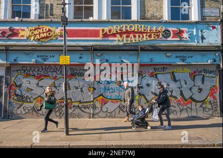 People walk past the site of The World Famous Gallery. Shopfront shutters are closed and covered in colourful graffiti. Portobello Road, London, UK Stock Photo
