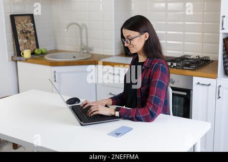 Cheerful young woman working on computer. Natural looking white female typing text on modern laptop keyboard in home kitchen Stock Photo