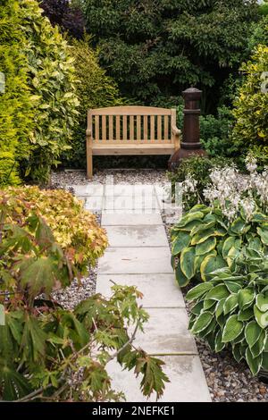A wooden garden bench and a chimenea in a leafy garden with a paved path leading in Stock Photo