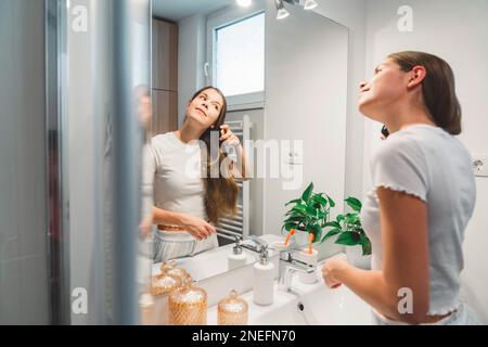 Side view of a woman brushing her long brown hair while looking at herself in the mirror  Stock Photo
