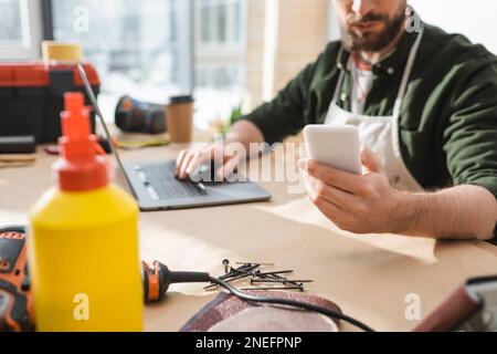 Cropped view of blurred carpenter using smartphone and laptop near tools on table,stock image Stock Photo