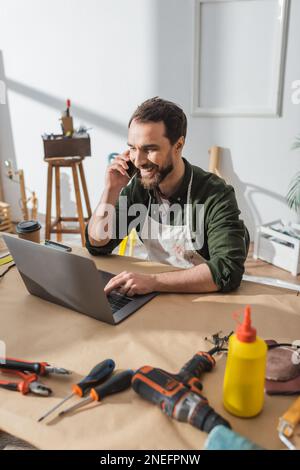 Cheerful carpenter talking on smartphone and using laptop near tools in workshop,stock image Stock Photo