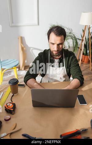 Bearded craftsman in apron using laptop near tools and coffee to go on table in workshop,stock image Stock Photo