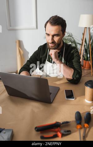 Craftsman in apron using laptop near tools and coffee to go in workshop,stock image Stock Photo