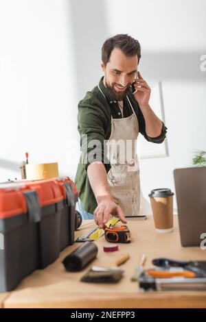Positive carpenter talking on smartphone and taking ruler in workshop,stock image Stock Photo