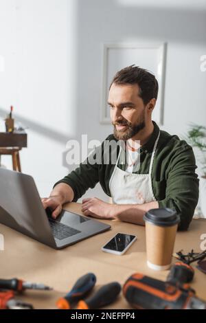 Smiling carpenter in apron using laptop near coffee and blurred tools in workshop,stock image Stock Photo