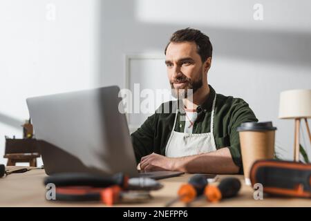 Bearded carpenter using laptop near blurred takeaway coffee and tools in workshop,stock image Stock Photo