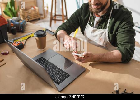 Cropped view of bearded craftsman using smartphone near laptop and coffee to go in workshop,stock image Stock Photo