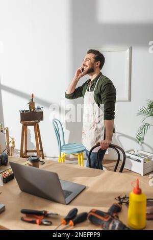 Positive craftsman talking on cellphone near tools and laptop on working table,stock image Stock Photo