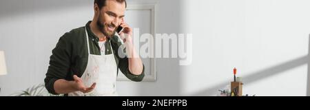 Smiling carpenter in apron talking on smartphone in workshop, banner,stock image Stock Photo