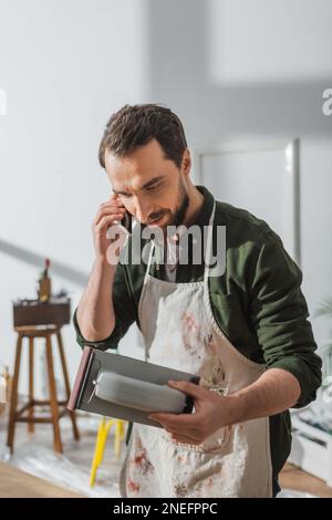 Carpenter in apron talking on smartphone and holding sandpaper in workshop,stock image Stock Photo
