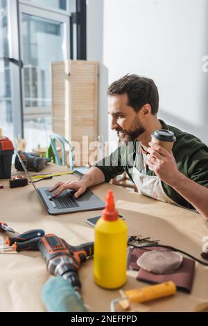 Positive bearded carpenter holding coffee to go and using laptop near tools in workshop,stock image Stock Photo