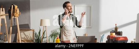 Smiling craftsman talking on cellphone near tools and laptop in studio, banner,stock image Stock Photo
