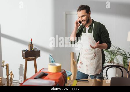 Smiling craftsman in apron talking on mobile phone near blurred toolbox in workshop,stock image Stock Photo