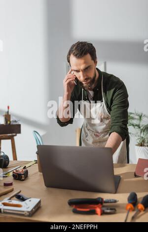 Bearded carpenter talking on smartphone and using laptop in workshop,stock image Stock Photo