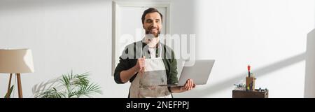 Cheerful carpenter in apron holding laptop and looking at camera in workshop, banner,stock image Stock Photo