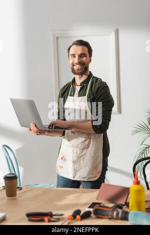Positive carpenter in apron using laptop near blurred tools and coffee in workshop,stock image Stock Photo