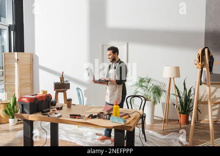Smiling carpenter in apron using laptop near tools and coffee in workshop,stock image Stock Photo