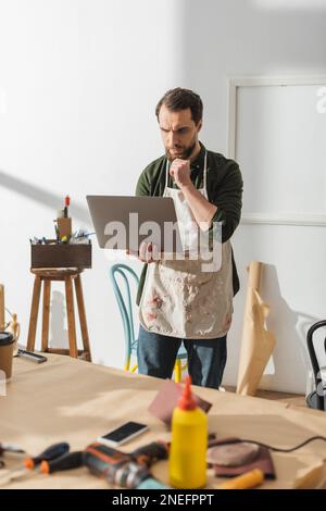 Pensive carpenter in apron using laptop in workshop,stock image Stock Photo
