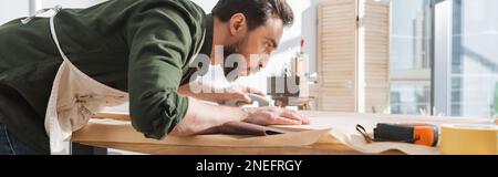 Side view of bearded carpenter blowing dust from wooden board in workshop, banner,stock image Stock Photo