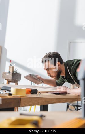 Brunette carpenter holding sandpaper and blowing on wooden board in workshop,stock image Stock Photo