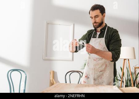 Bearded carpenter in apron using calipers while working with wooden board in workshop,stock image Stock Photo