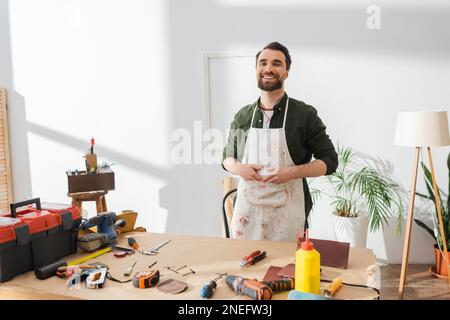 Positive restorer in apron looking at camera near tools on table in workshop,stock image Stock Photo
