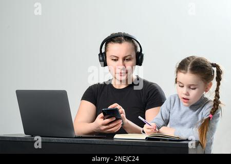 a young woman in headphones works on a laptop and at the same time exercises with her daughter, mother and daughter interaction. Stock Photo