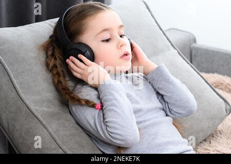 Little girl lies on the sofa and listens to music in headphones and rests, copy space and home rest of the child, children's relaxation with music Stock Photo