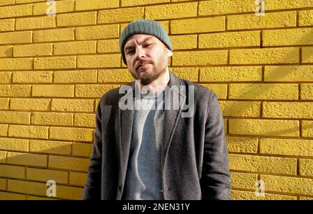 Stylish urban adult man wearing hat and coat standing against yellow brick wall and looking at camera. Stock Photo