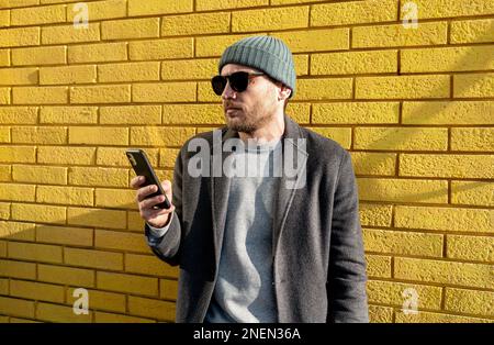 Stylish urban man in hat with sunglasses standing against yellow brick wall and using his cell phone. Stock Photo