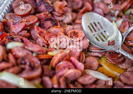 Sliced smoked fried Kielbasa calabrese sausage with onion as Brazilian Linguica Bebada appetizer dish and serving spoon in buffet tray for catered eve Stock Photo