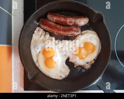 Double Fried Eggs Sunny Side Up, fried sausages on a cast iron frying pan on a cooktop background Stock Photo