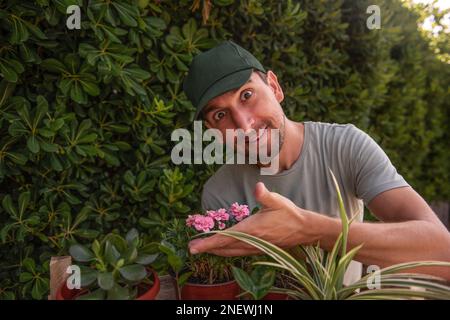 Man gardener in green cap smiles as smells indoor plants in front of living evergreen fence Phillyrea latifolia. Allergy to flowers. Natural holiday g Stock Photo