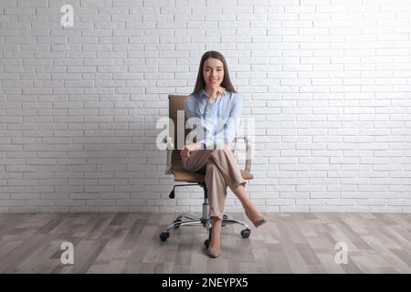 Young businesswoman sitting in office chair near white brick wall indoors Stock Photo