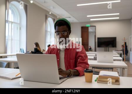 Puzzled student black hipster man sits at table with textbooks laptop studying in lecture classroom. Stock Photo