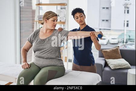 Try keeping your arm outstretched for as long as possible. a young physiotherapist helping her mature patient exercise with dumbbells at a clinic. Stock Photo