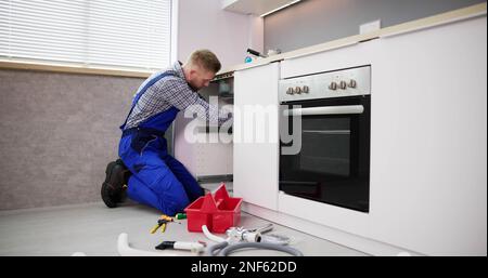 Man Looking At Male Plumber Cleaning Clogged Sink Pipe With Drained Cable In Kitchen Stock Photo