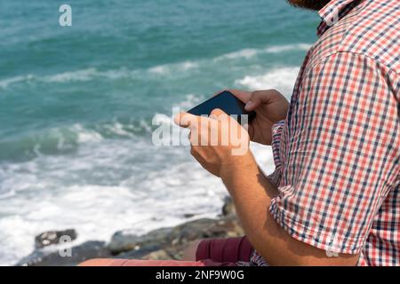 Man in a plaid shirt sitting on the seashore on a sunny day and holding a mobile phone in his hands Stock Photo