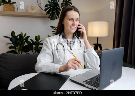 Smiling cute young female doctor physician holding cell phone talking on mobile, using laptop. Healthcare professional answering call giving remote co Stock Photo