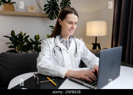 Smiling young woman doctor or nurse in white medical coat and stethoscope using laptop computer. Happy caucasian female GP work online consult patient Stock Photo