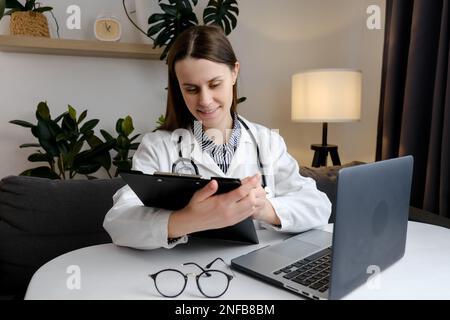 Close up of smiling doctor woman filling paper medical records. Practitioner in white coat doing paperwork at workplace with laptop, writing notes, pr Stock Photo